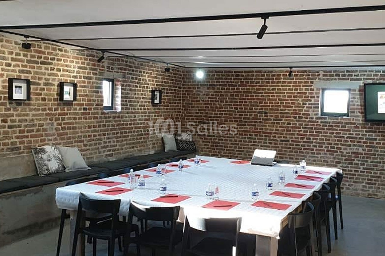 Salle de réunion avec murs en briques, table rectangulaire dressée avec documents rouges, chaises noires et lumière…