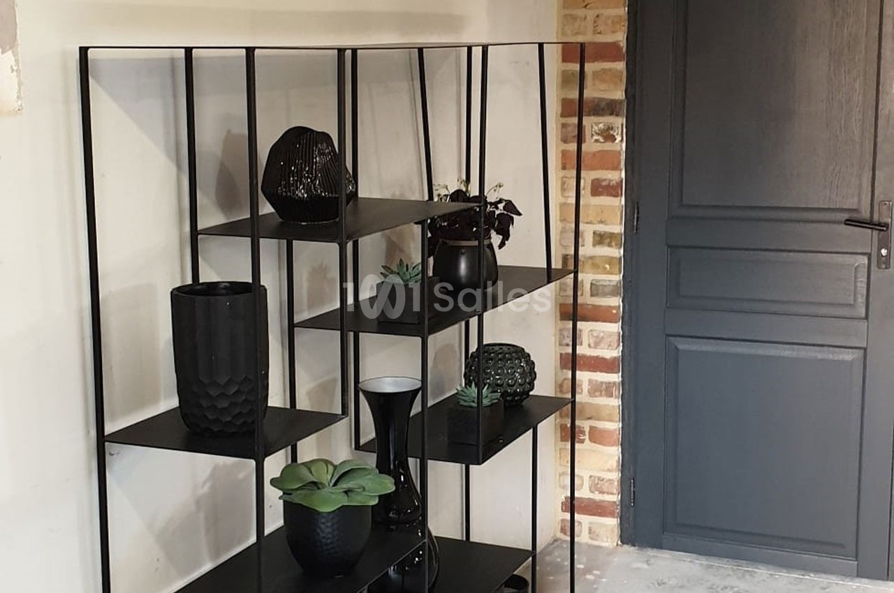 Étagère noire moderne avec des objets décoratifs et des plantes, placée près d'un mur en brique et d'une porte grise.