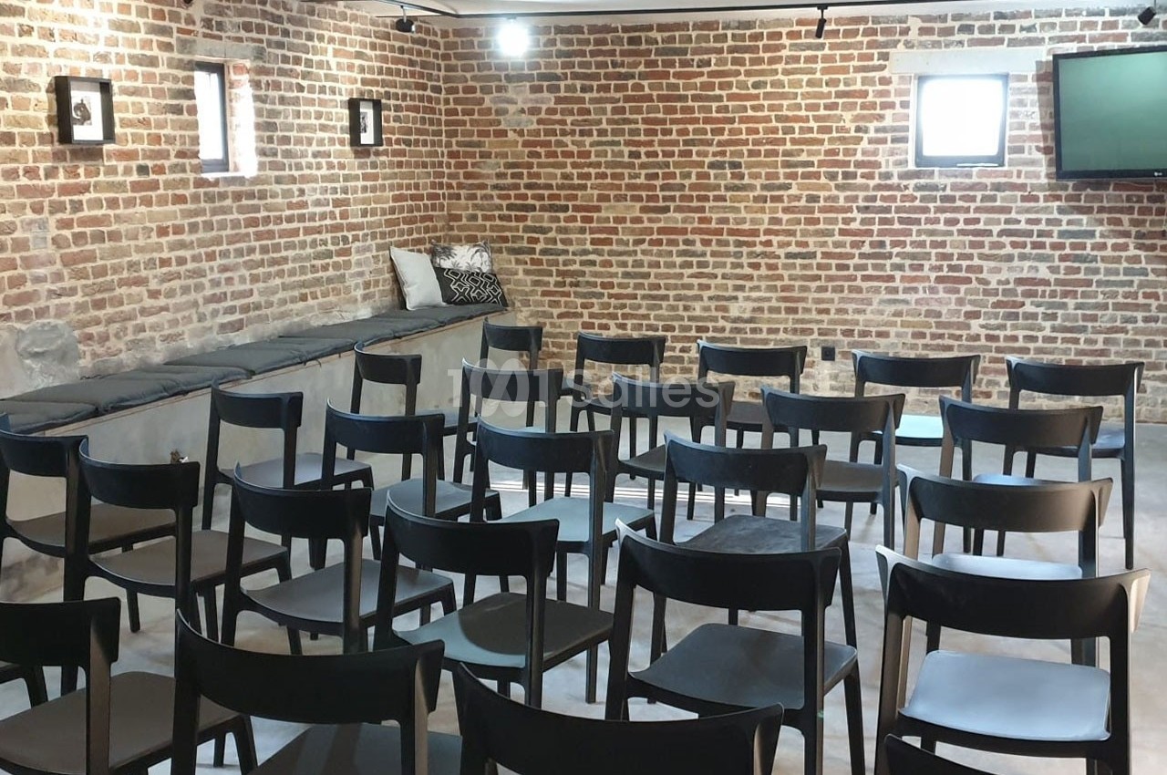 Salle avec des murs en briques, rangées de chaises noires alignées et un écran fixé au mur.