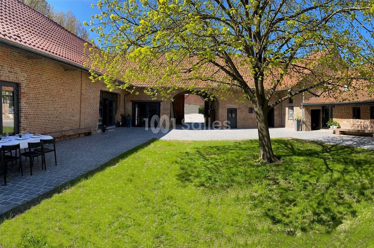 Cour intérieure d'une ferme rénovée en briques, avec un arbre au centre et des tables dressées sur la gauche.
