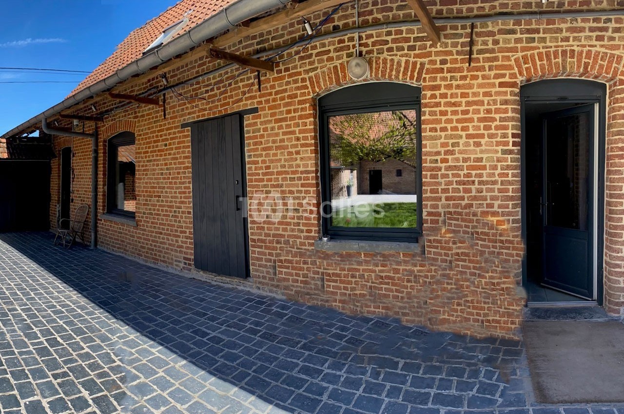 Façade en briques rouges avec portes et fenêtres noires, donnant sur une cour pavée ensoleillée.