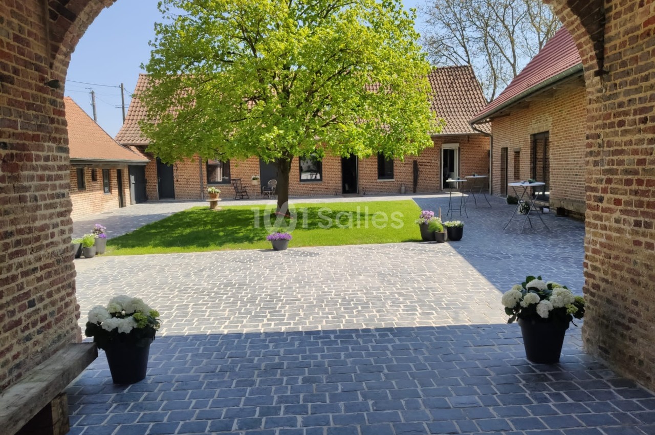 Cour pavée avec un arbre central, entourée de bâtiments en briques, pots de fleurs et mobilier de jardin.