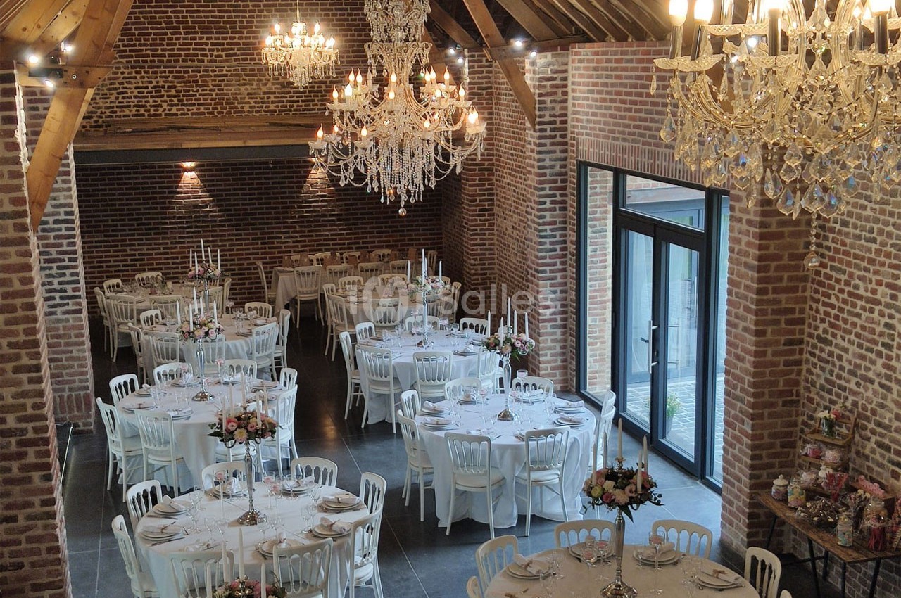 Salle de réception en briques avec tables rondes dressées, chaises blanches et lustres en cristal suspendus.