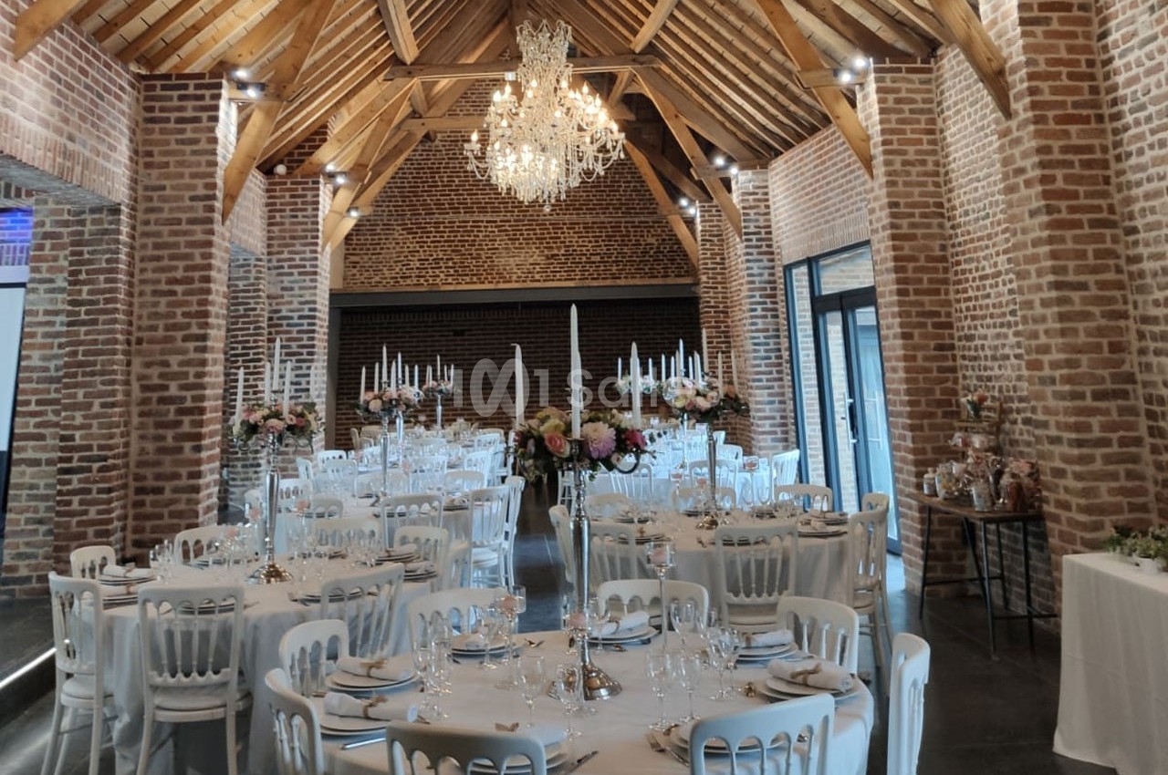 Salle de réception avec murs en briques, poutres apparentes, tables rondes dressées et lustre central.