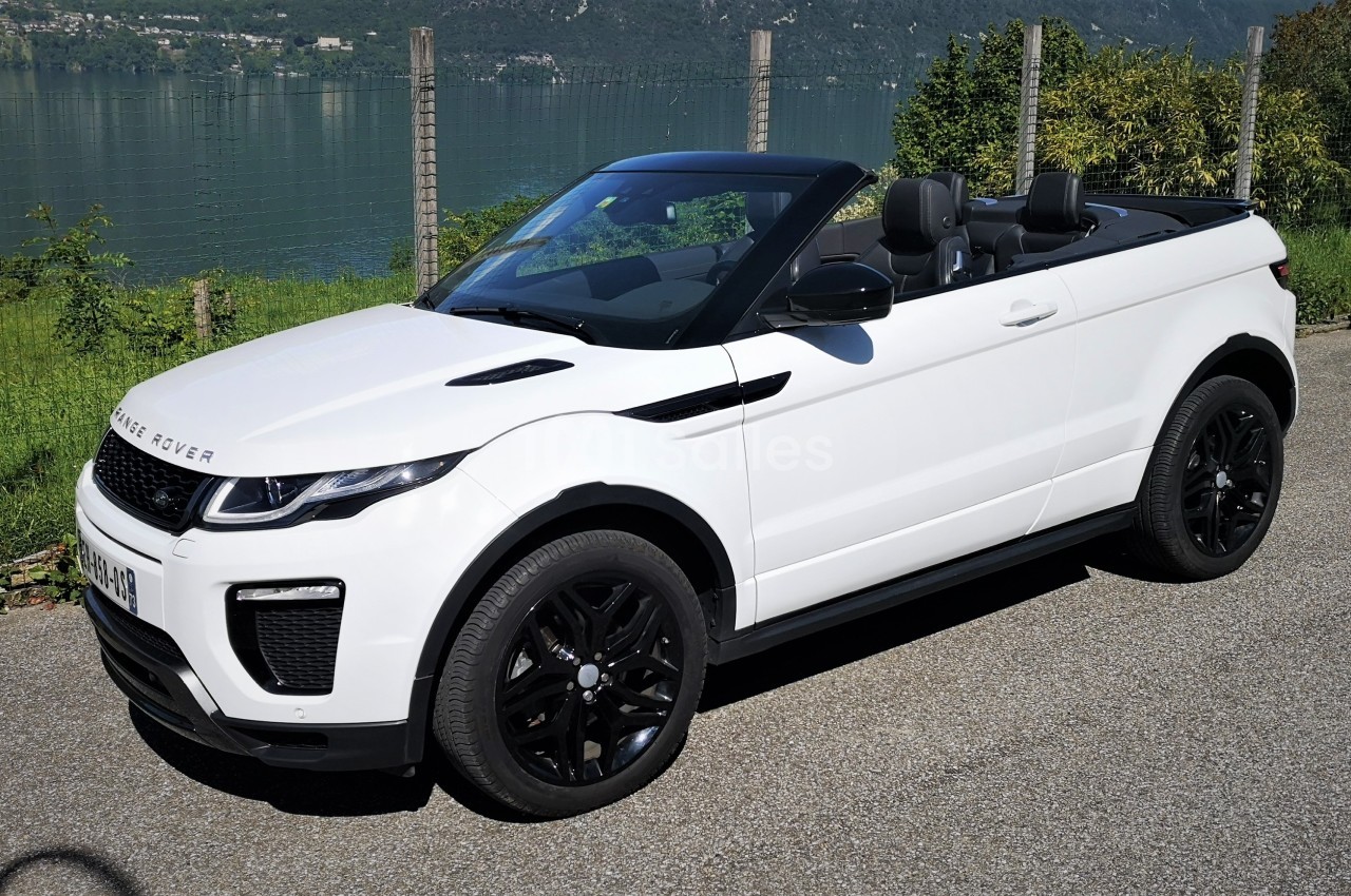 Voiture cabriolet blanche stationnée près d'un lac, avec des collines en arrière-plan.