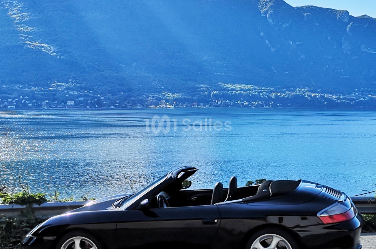 Voiture décapotable noire garée près d'un lac, avec des montagnes et un ciel bleu en arrière-plan.