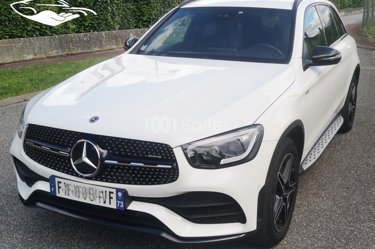SUV blanc Mercedes-Benz stationné sur une route, avec une plaque d'immatriculation visible et des haies en arrière-plan.