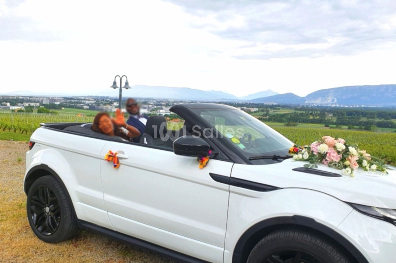 Une voiture décapotable blanche décorée de fleurs, garée dans un paysage de vignobles avec des montagnes en arrière-plan.