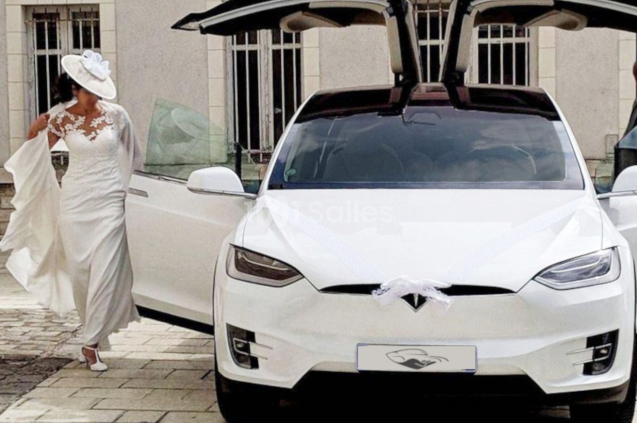Une mariée en robe blanche sort d'une voiture électrique blanche décorée pour un mariage, avec des portes papillon ouvertes.