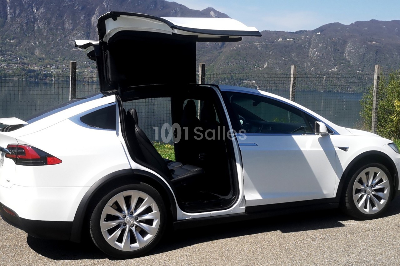 Voiture blanche avec portes arrière ouvertes en ailes de mouette, stationnée près d'un lac et de montagnes.