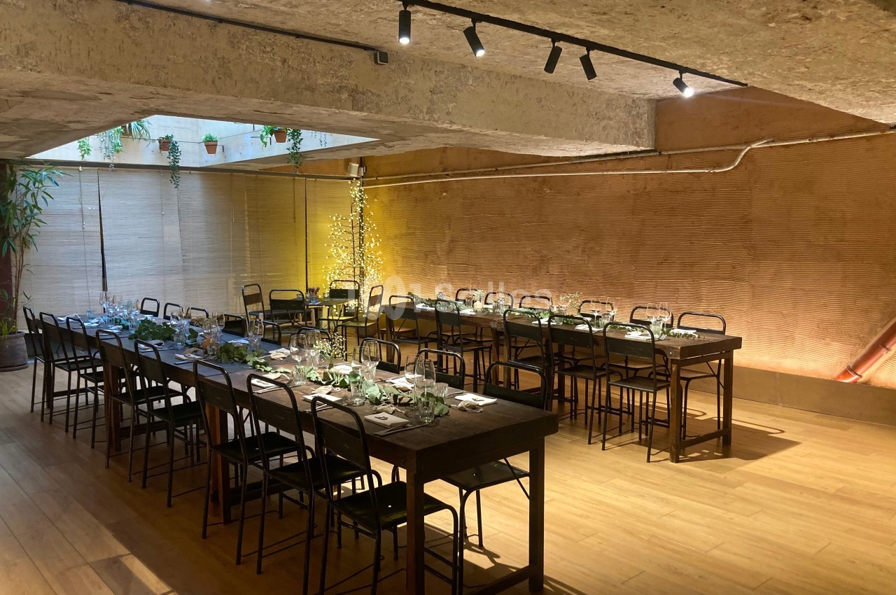 Salle avec deux longues tables dressées, éclairage chaleureux, murs en pierre et plantes suspendues.