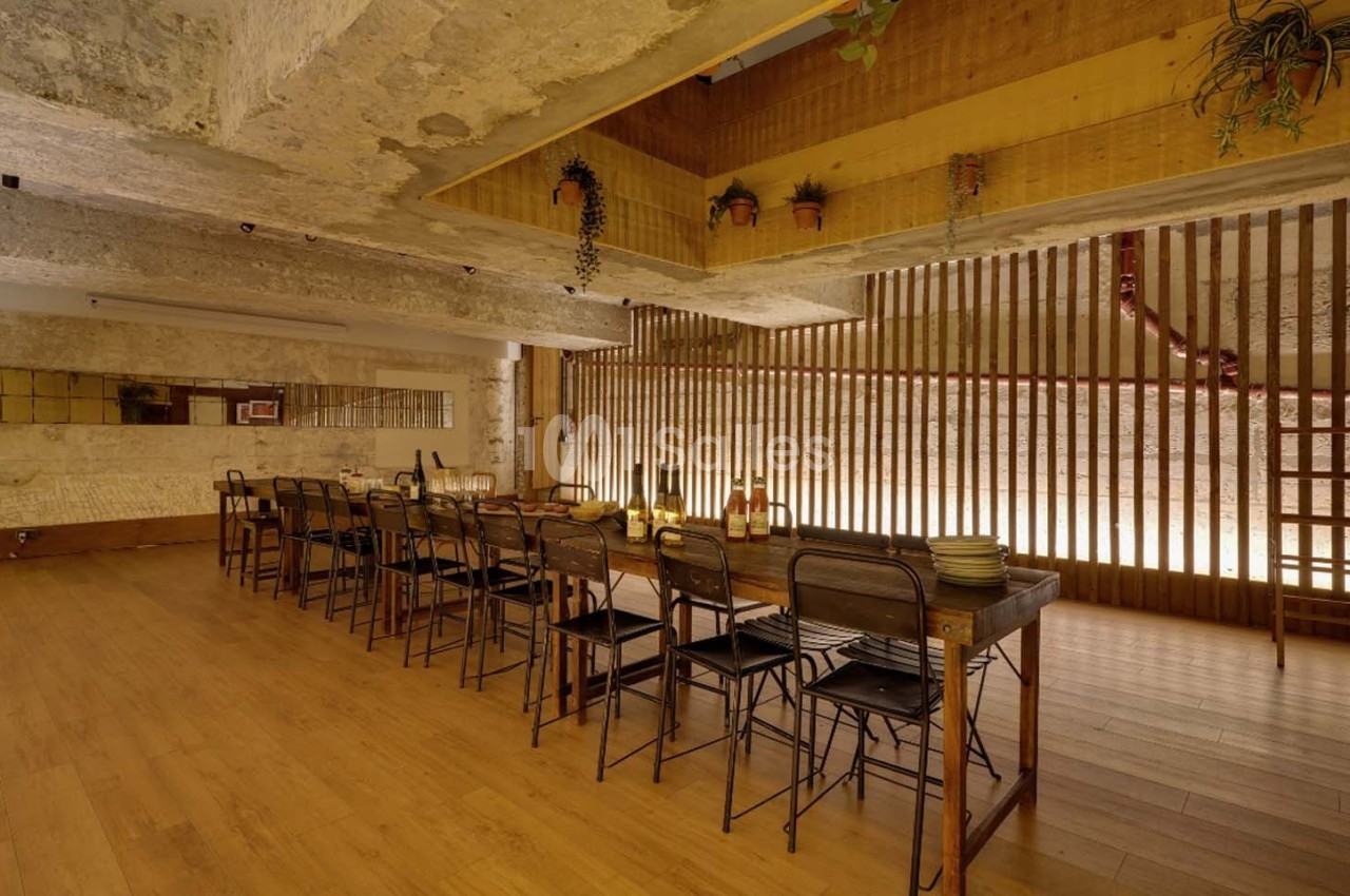 Salle à manger avec une grande table en bois, des chaises métalliques, un éclairage chaleureux et des plantes suspendues.