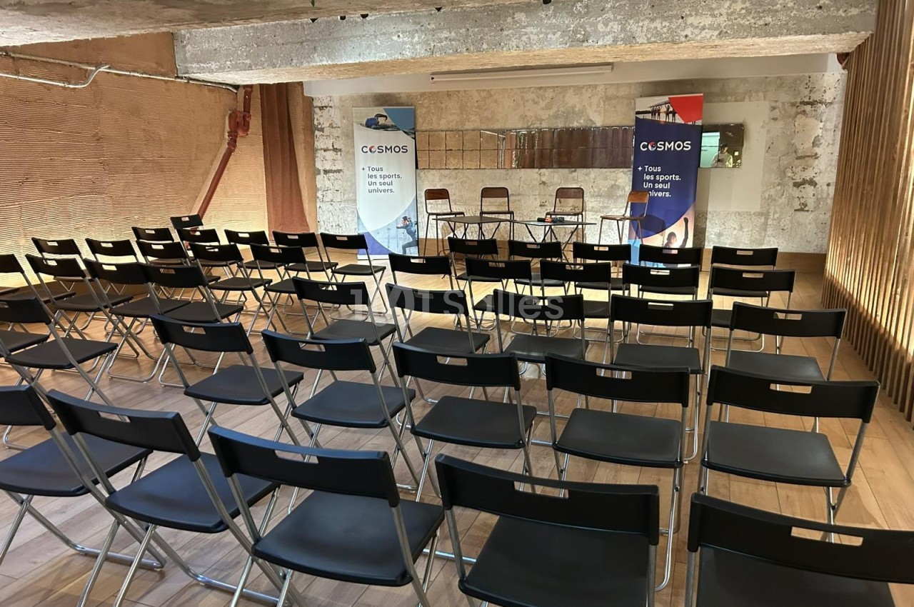 Salle de conférence vide avec des rangées de chaises noires pliantes et deux kakémonos colorés en arrière-plan.