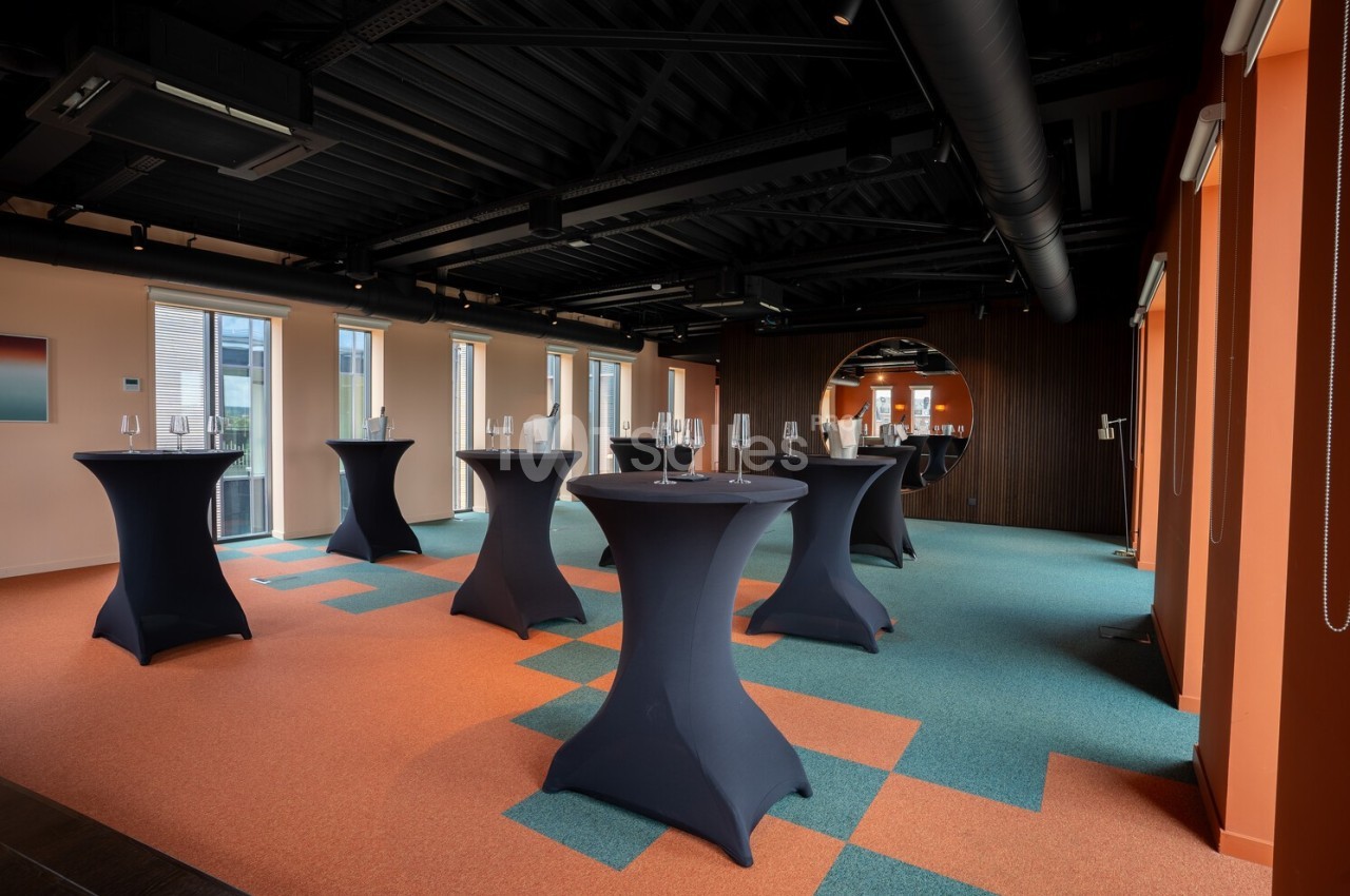 Salle moderne avec tables hautes noires, sol coloré orange et bleu, grandes fenêtres et éclairage naturel.