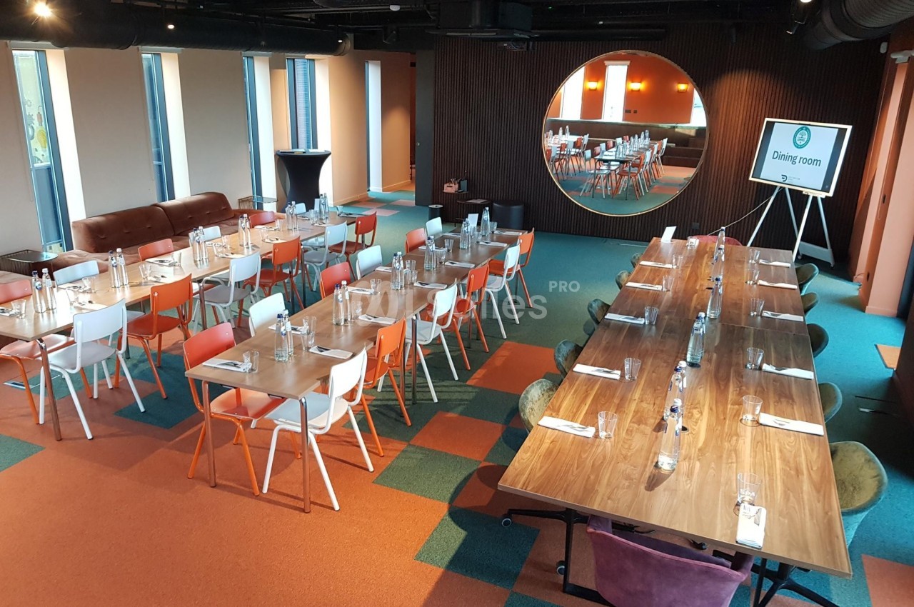 Salle de réunion moderne avec tables en bois, chaises colorées et miroir rond reflétant l'espace.