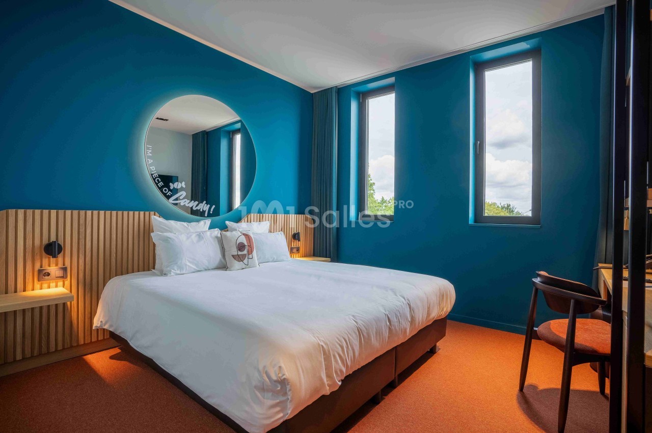 Chambre d'hôtel moderne avec murs bleus, lit double, miroir rond, deux fenêtres et éclairage chaleureux.