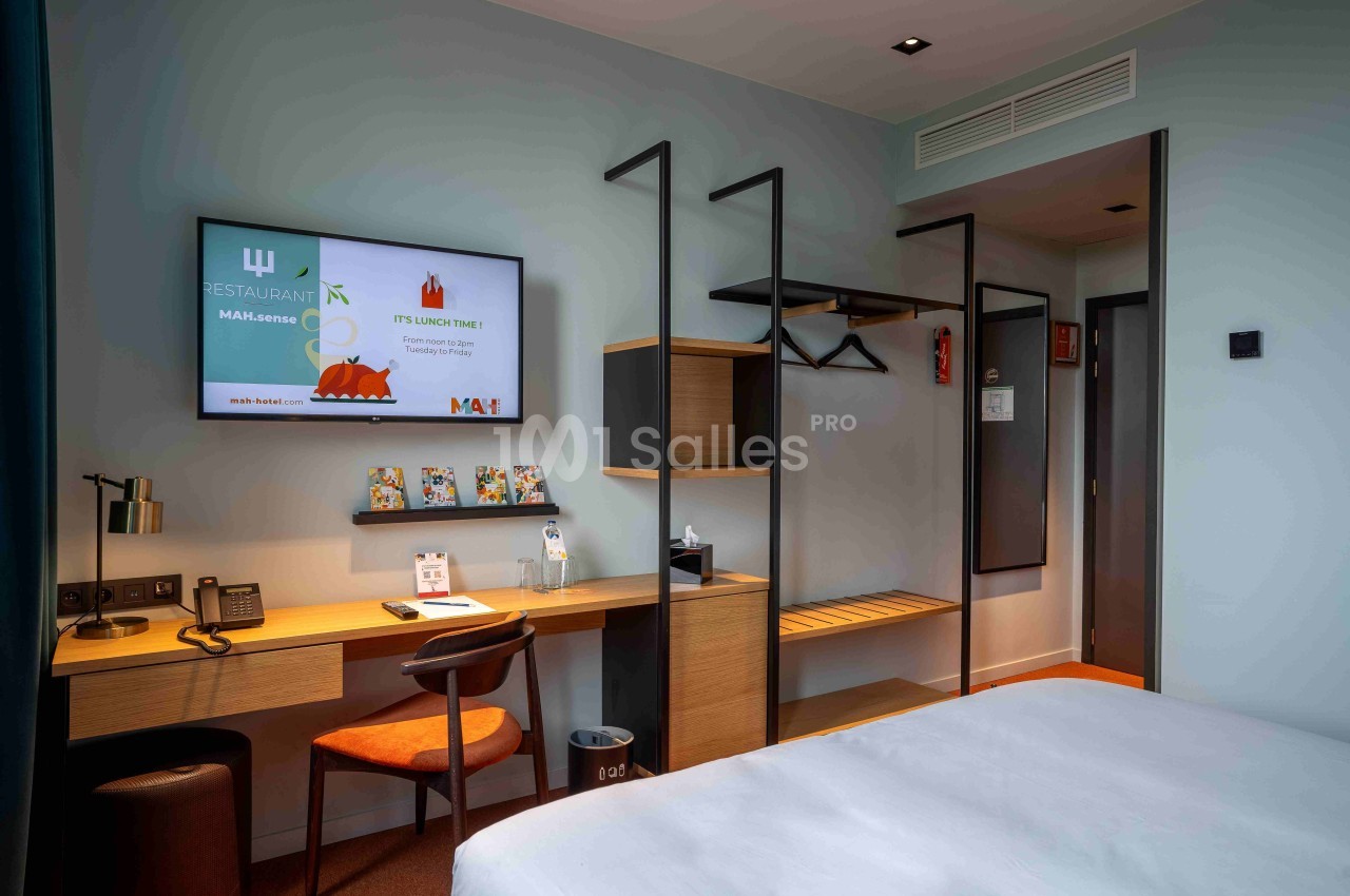 Chambre d'hôtel moderne avec bureau, téléviseur mural, étagères ouvertes et lit partiellement visible au premier plan.