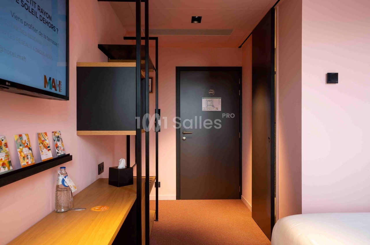 Chambre d'hôtel avec murs roses, bureau en bois, étagères, télévision murale et porte d'entrée noire.