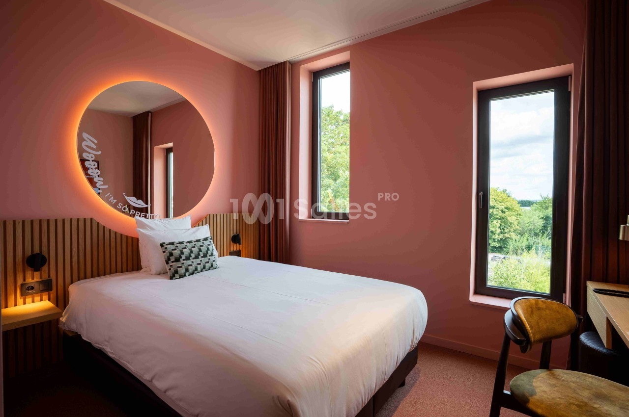 Chambre d'hôtel avec lit double, mur rose, miroir rond éclairé, bureau, chaise et fenêtres donnant sur un paysage verdoyant.