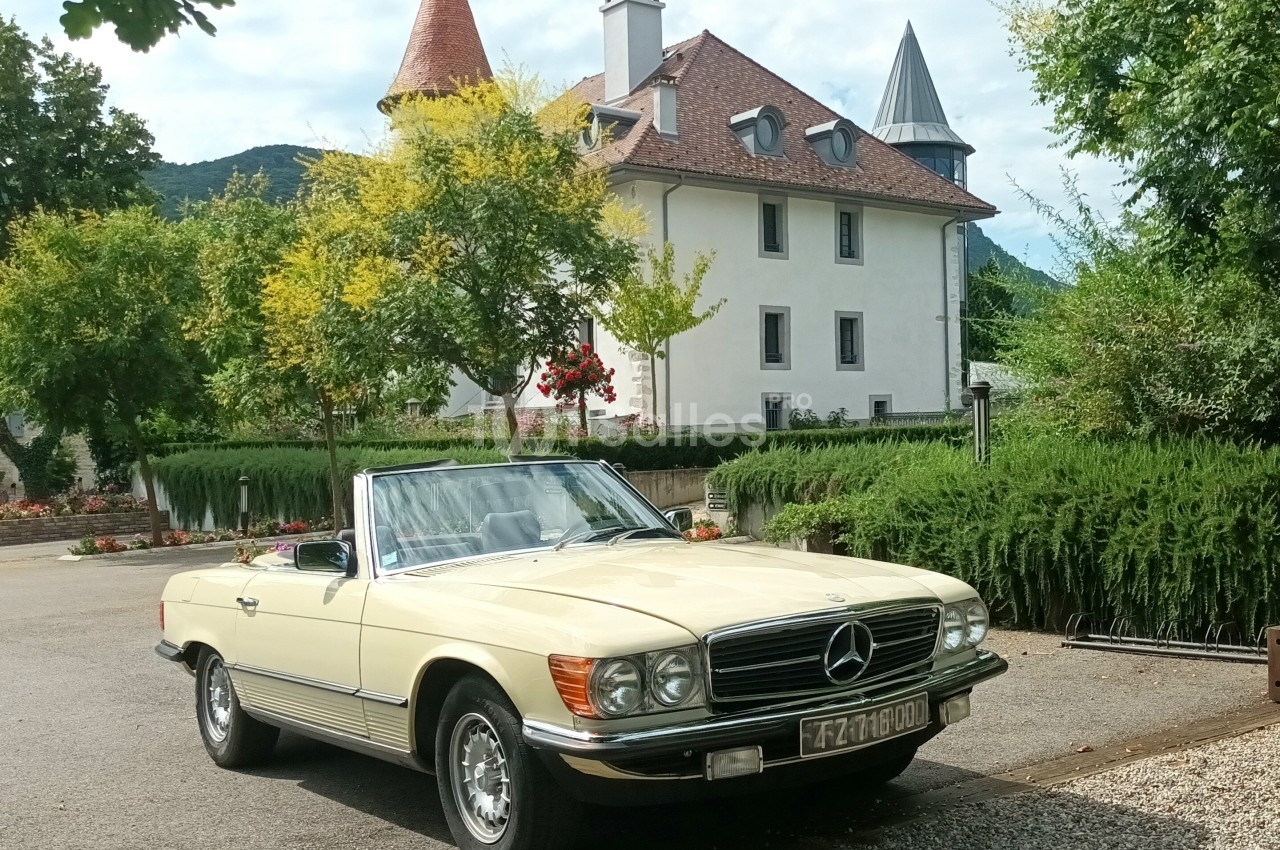 Voiture décapotable Mercedes ancienne beige stationnée devant un bâtiment à toit en tuiles entouré de verdure.