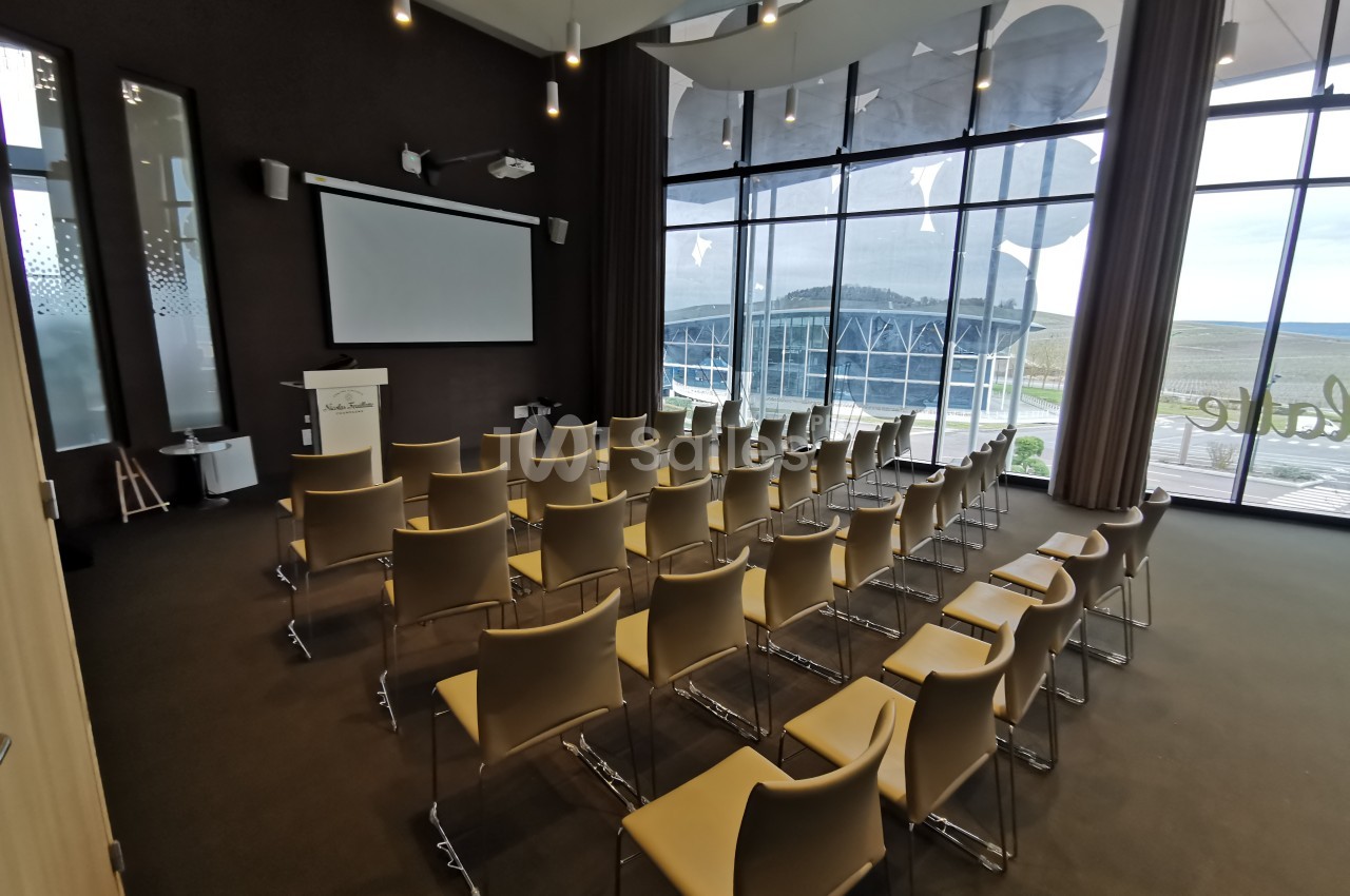 Salle de conférence moderne avec chaises alignées, écran de projection et grandes baies vitrées donnant sur l'extérieur.