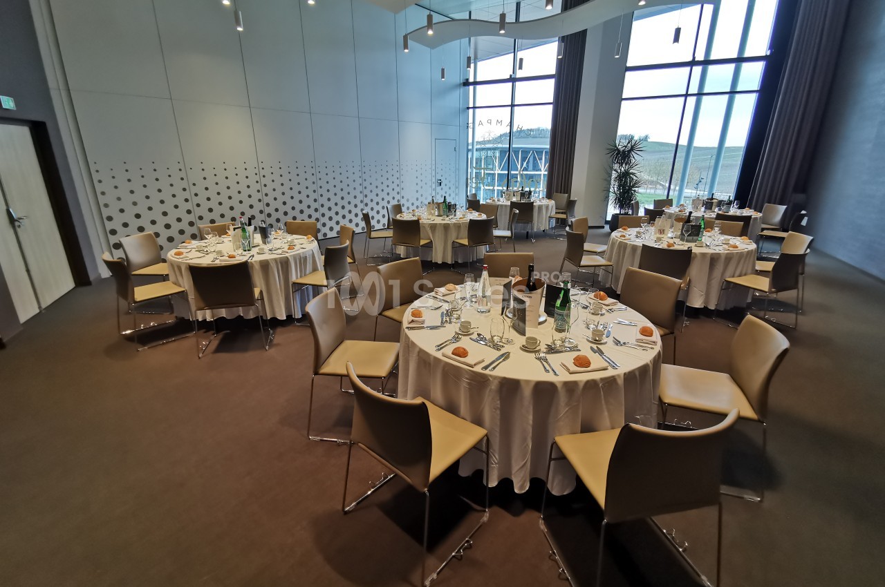 Salle de réception avec tables rondes dressées pour un repas, entourées de chaises, près de grandes baies vitrées.