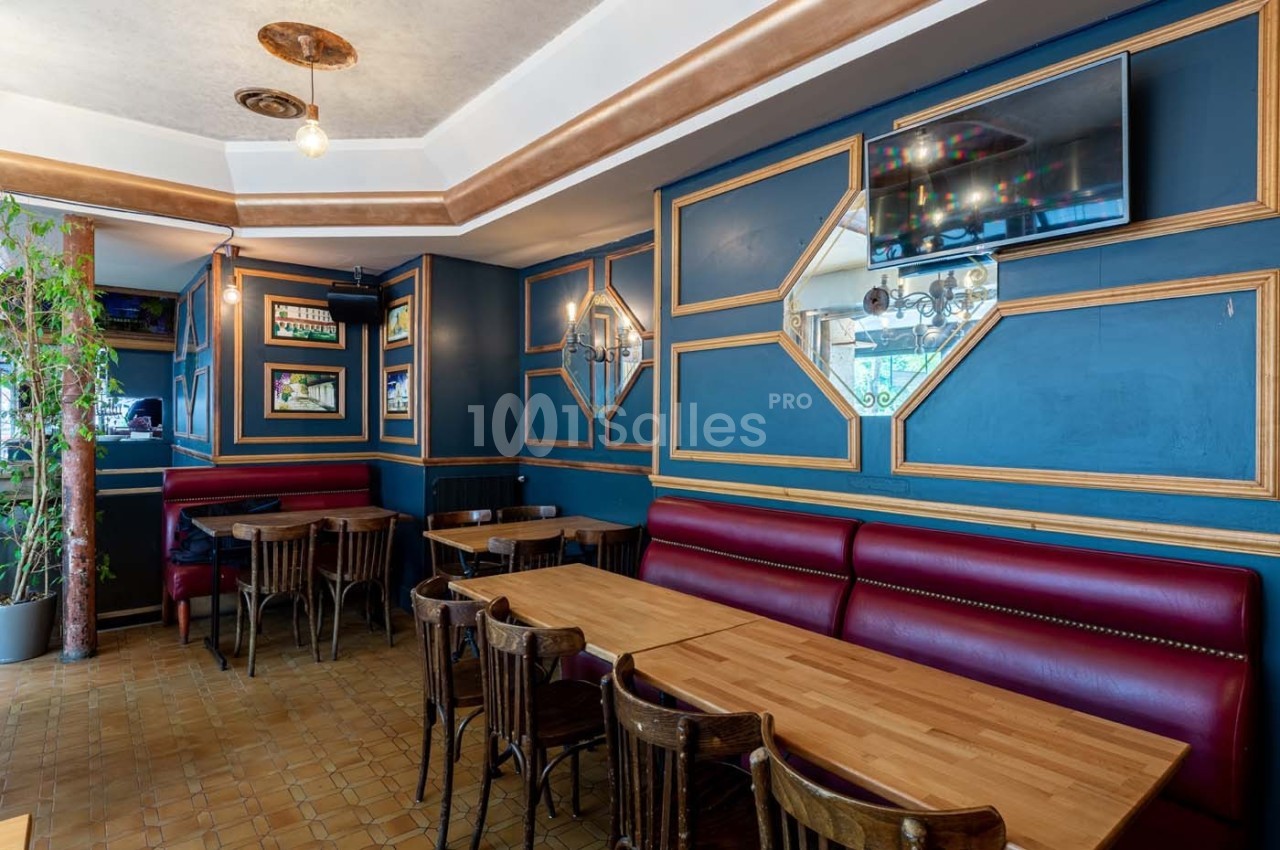 Salle de restaurant avec tables en bois, banquettes rouges, murs bleus ornés de cadres dorés et une télévision fixée.