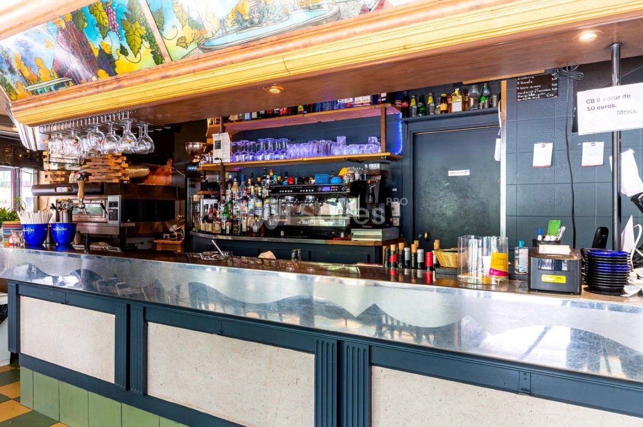 Comptoir de bar avec étagères garnies de verres, bouteilles et machine à café, dans un décor coloré et lumineux.