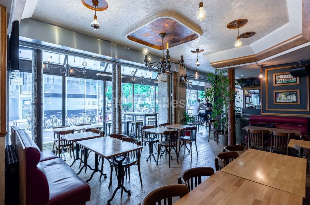 Salle de restaurant lumineuse avec tables en bois, chaises assorties et grandes baies vitrées donnant sur la rue.
