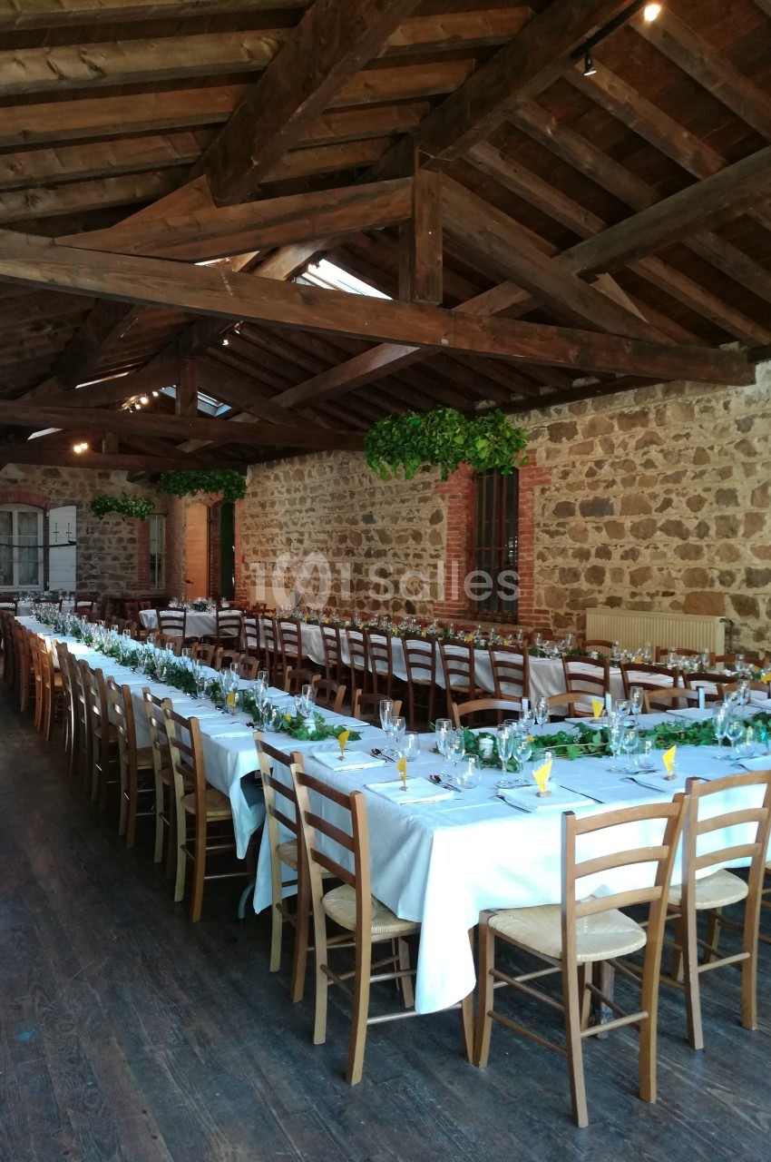 Salle en pierre avec plafond en bois, tables longues dressées avec nappes blanches, chaises en bois et décoration végétale.