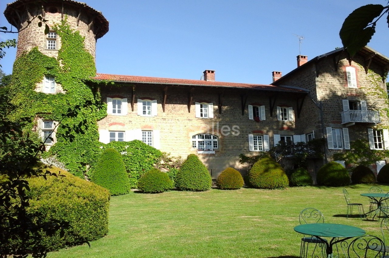Façade d'un bâtiment en pierre avec une tour, entouré de verdure et d'un jardin aménagé avec tables en fer forgé.