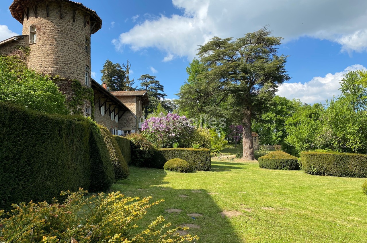 Jardin verdoyant avec pelouse, arbustes taillés, arbres imposants et bâtiment en pierre avec tour ronde sous un ciel bleu.