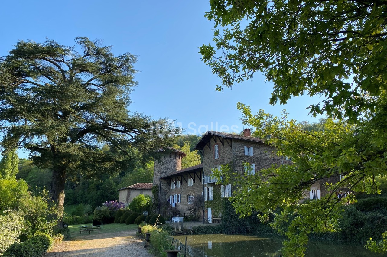 Grande maison en pierre entourée de verdure, avec un étang au premier plan et un grand arbre sur la gauche.