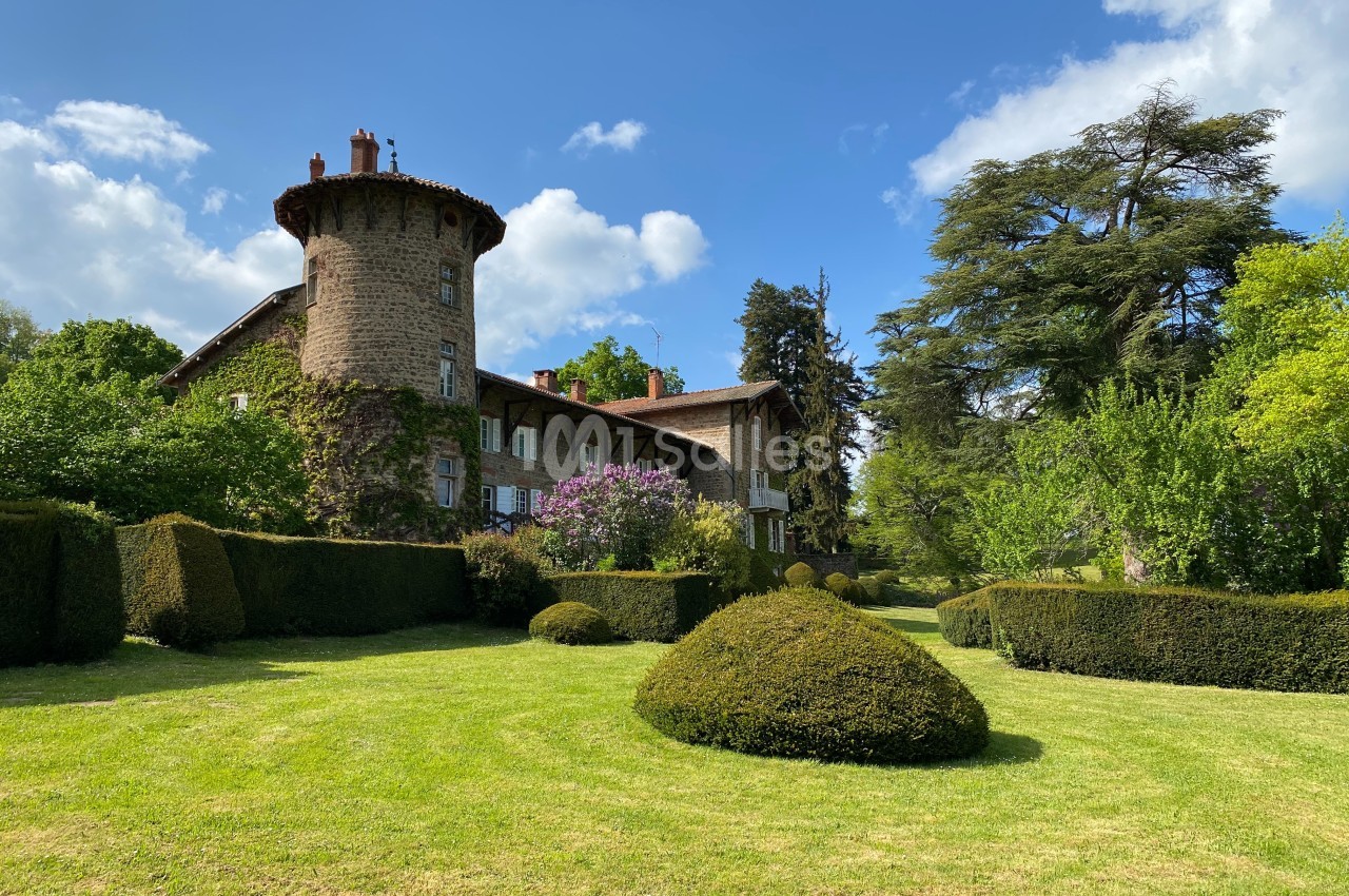 Grande maison en pierre avec une tour ronde, entourée de haies taillées, d'arbres et d'une pelouse sous un ciel bleu.