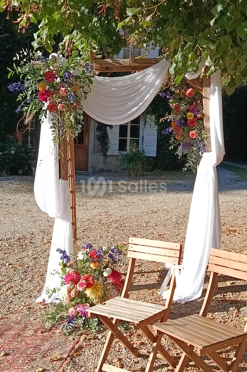 Arche de mariage en bois décorée de fleurs colorées et de voilages blancs, avec deux chaises en bois devant.