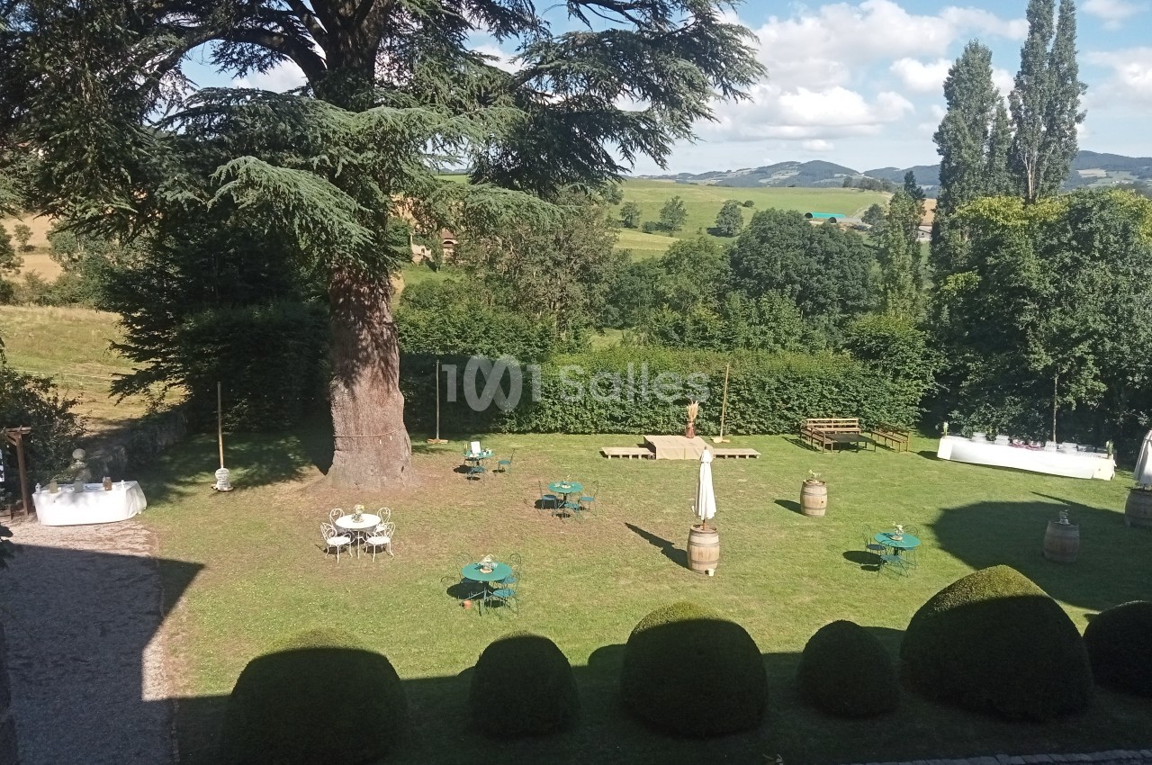 Jardin avec pelouse, tables et parasols, entouré de haies et d'arbres, avec vue sur une campagne vallonnée.