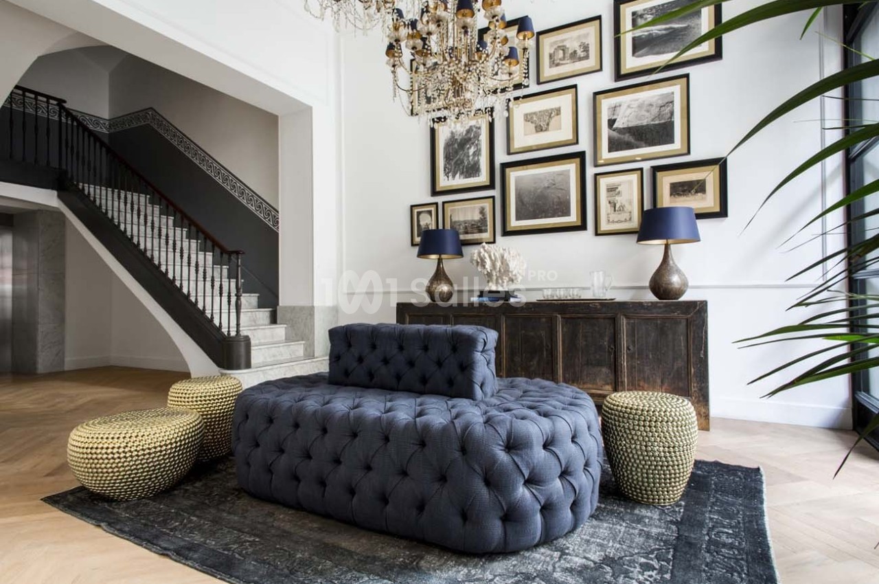 Salon élégant avec canapé capitonné gris, poufs tressés, tapis sombre, buffet en bois, cadres au mur et lustre imposant.