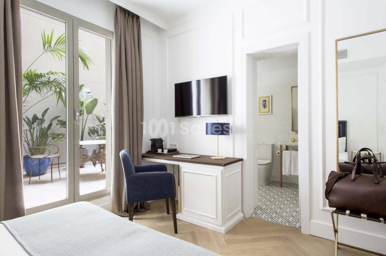 Chambre lumineuse avec bureau, fauteuil bleu, télévision murale, salle de bain attenante et accès à une terrasse avec…