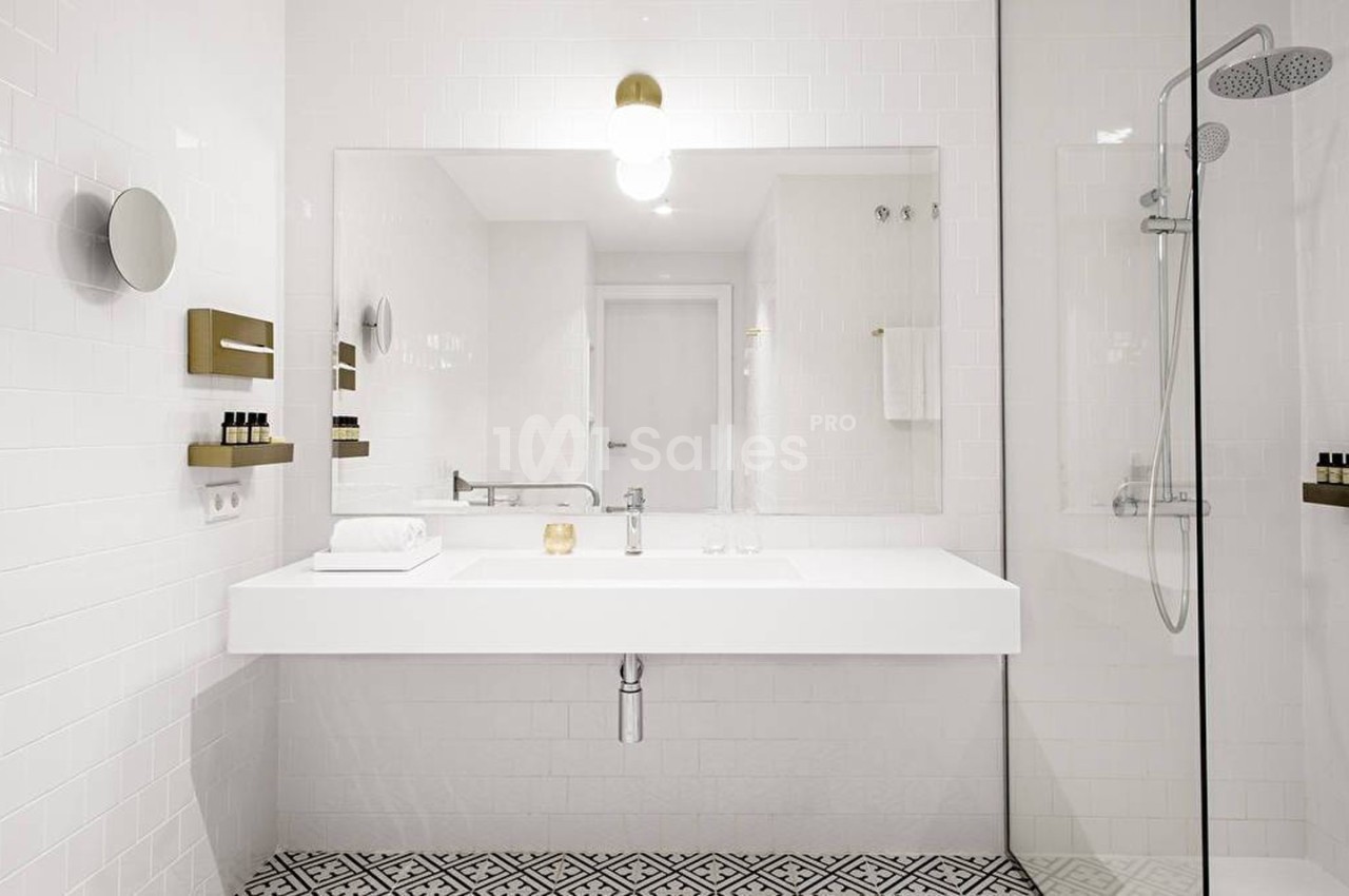 Salle de bain moderne avec lavabo blanc, miroir mural, douche vitrée et carrelage au sol à motifs géométriques.