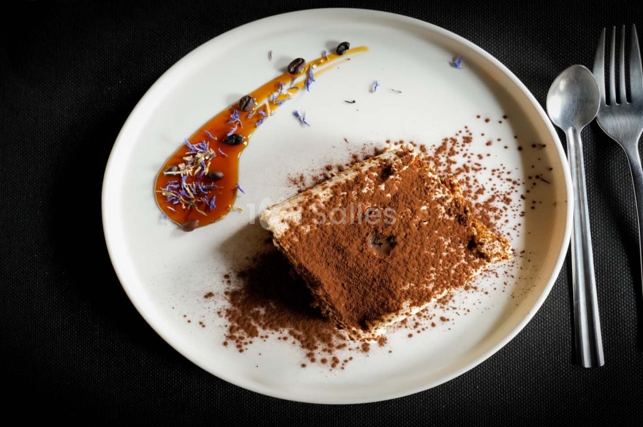 Part de tiramisu saupoudrée de cacao, servie avec une décoration de caramel et fleurs comestibles sur une assiette blanche.
