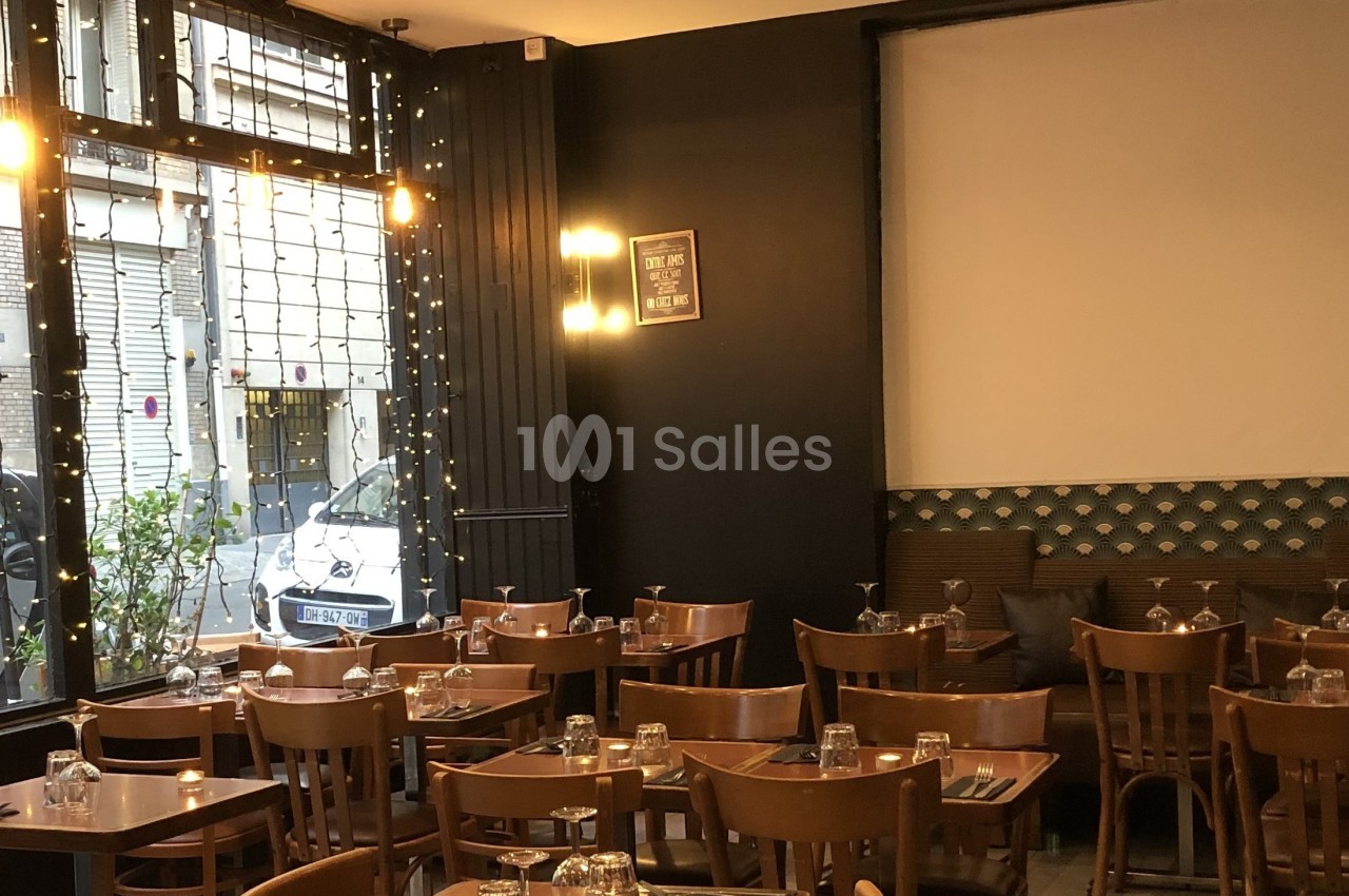 Salle de restaurant avec tables en bois, chaises assorties, grandes fenêtres et guirlandes lumineuses.