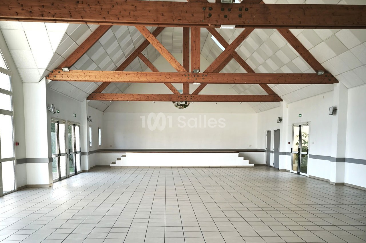 Grande salle vide avec carrelage au sol, poutres apparentes au plafond et larges fenêtres laissant entrer la lumière.