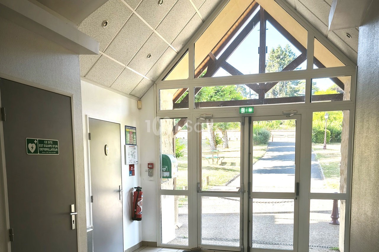 Entrée vitrée d'un bâtiment avec porte double, panneau de sortie de secours et vue sur un chemin extérieur arboré.