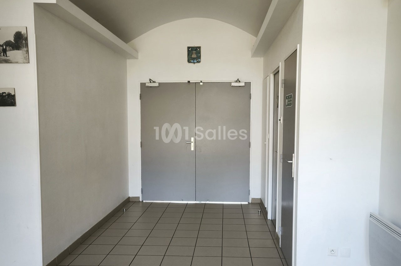 Porte double grise dans un couloir lumineux avec murs blancs, sol carrelé et panneaux indiquant des toilettes.