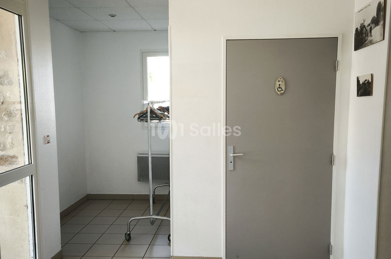 Porte grise avec pictogramme, portant à vêtements sur roulettes et fenêtre dans un couloir carrelé.