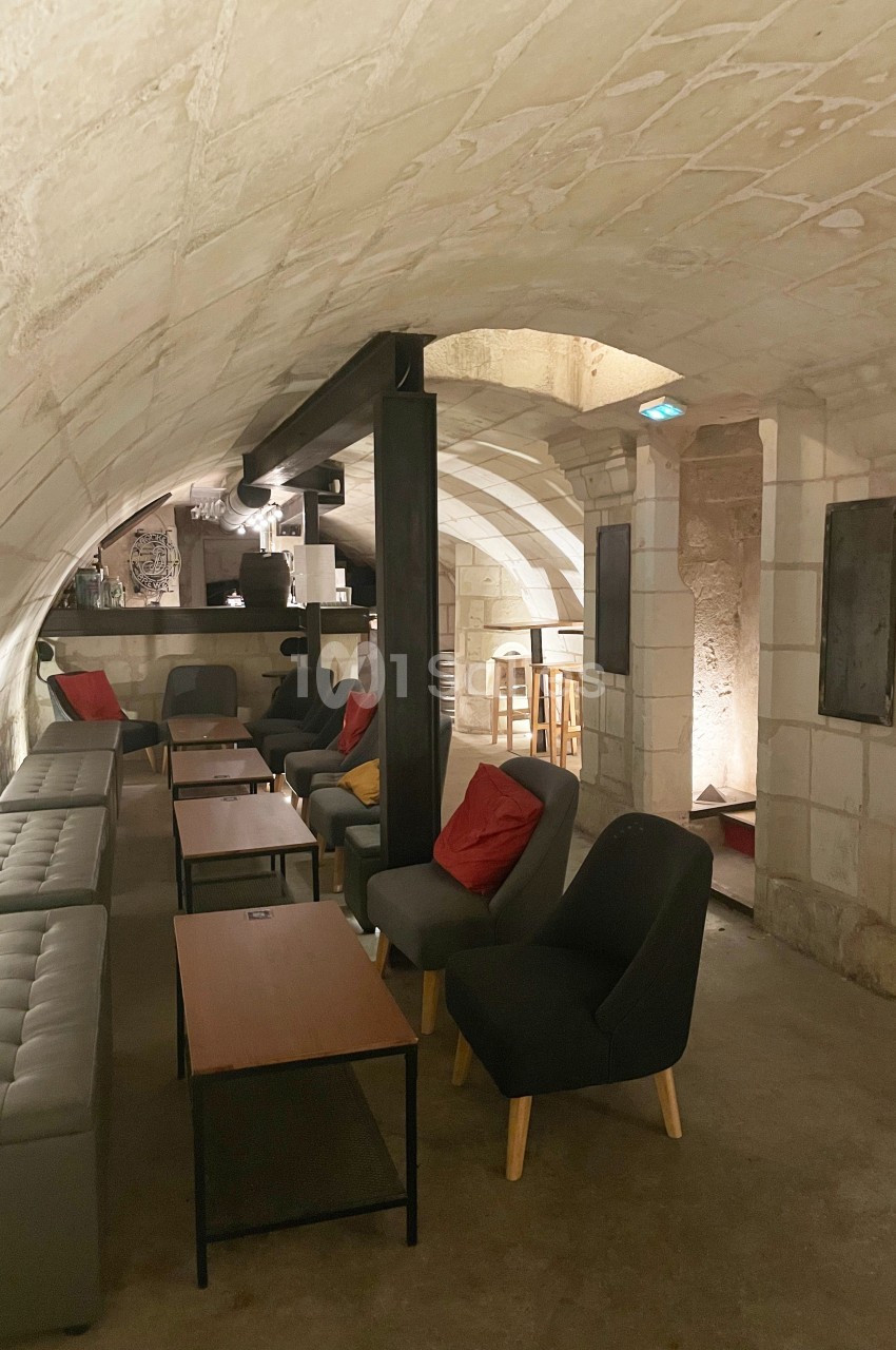 Salle voûtée en pierre avec canapés, fauteuils, tables basses et un bar en arrière-plan.