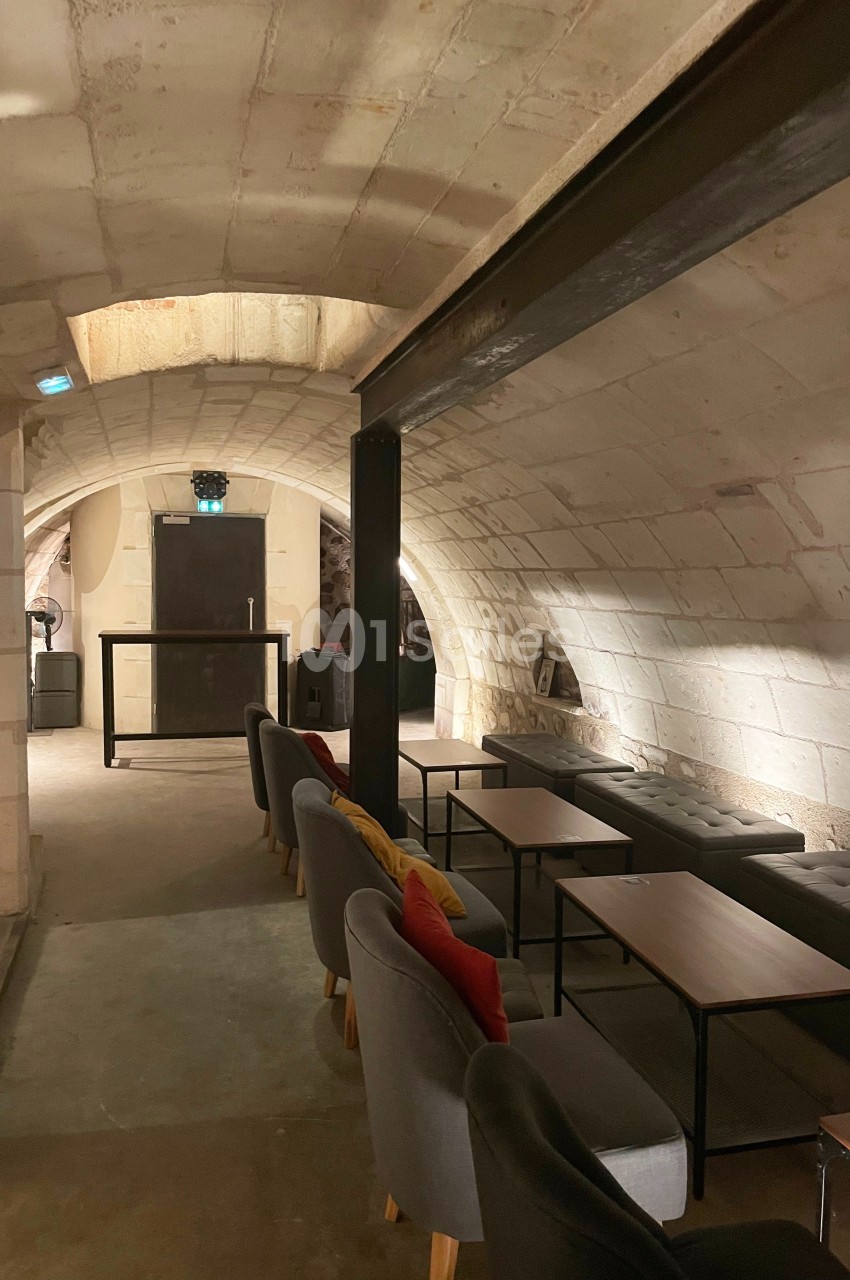 Salle voûtée en pierre avec des tables, des fauteuils gris et une lumière tamisée.