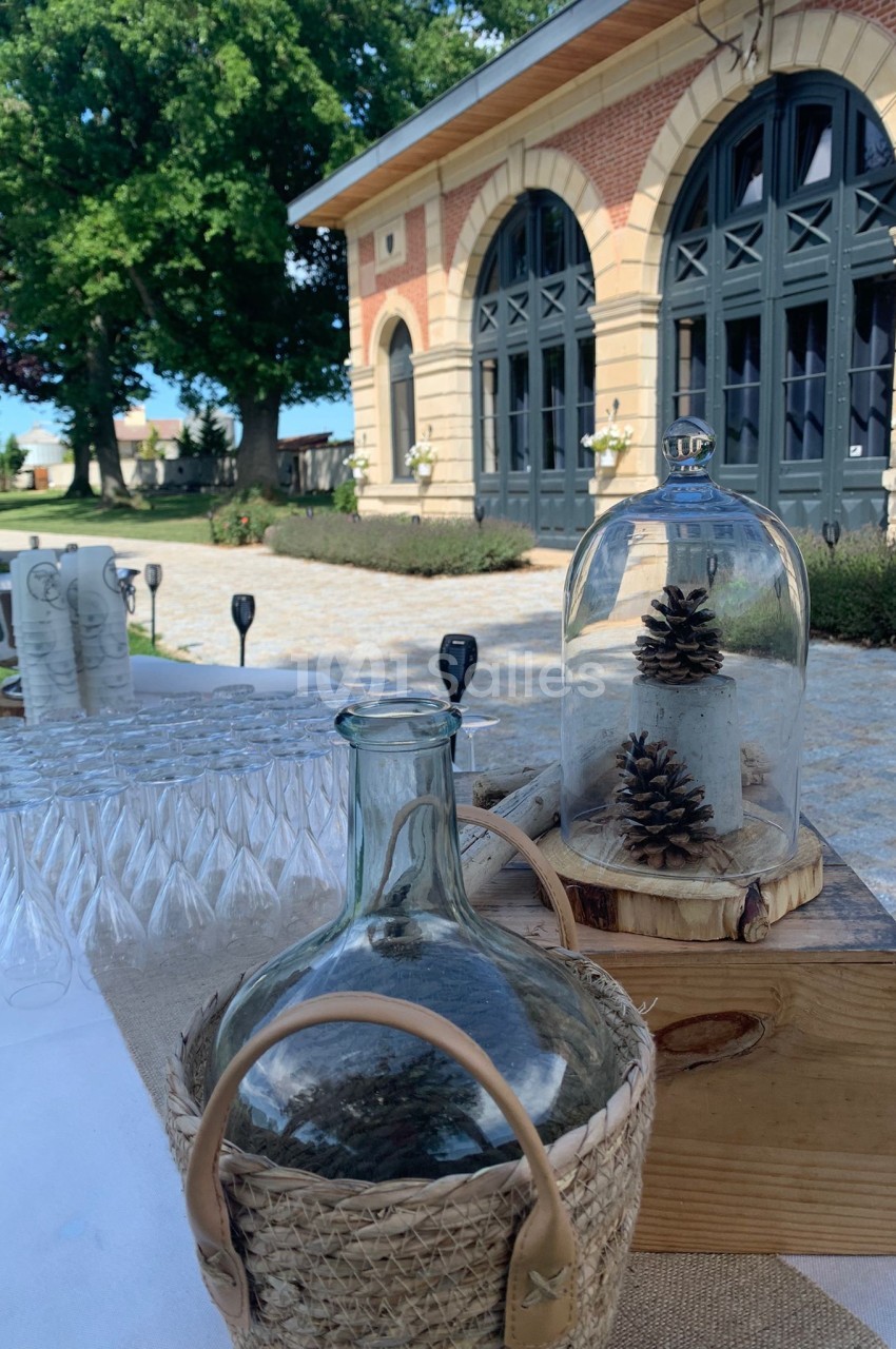 Table décorée en extérieur avec des verres, une bonbonne en verre et des pommes de pin sous cloche devant un bâtiment en…
