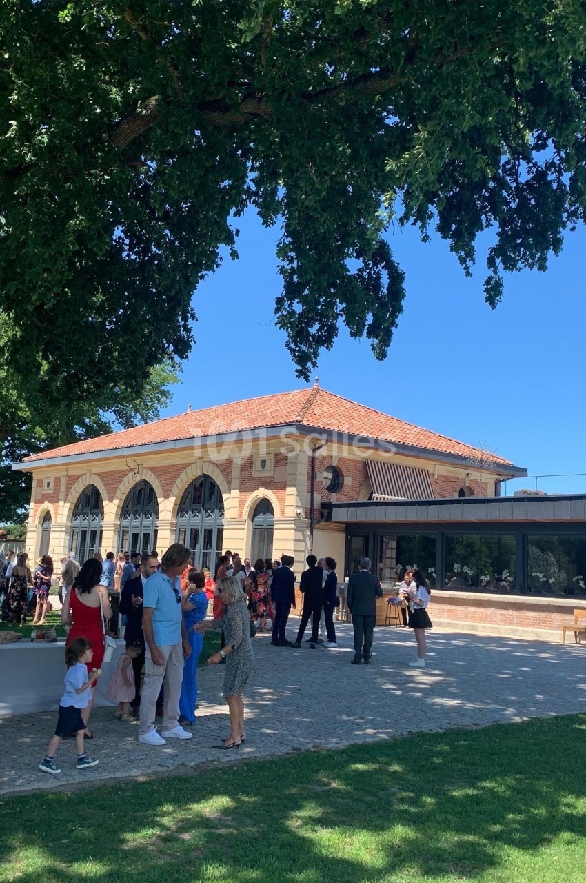 Groupe de personnes rassemblées devant un bâtiment en briques avec de grandes arches, sous un ciel bleu et des arbres.