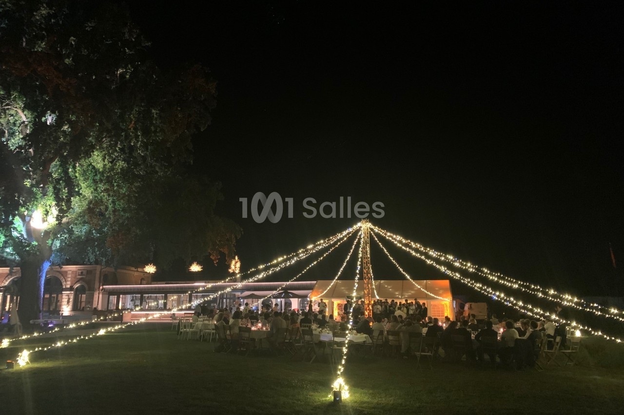 Tente éclairée par des guirlandes lumineuses dans un jardin, entourée de convives en soirée.